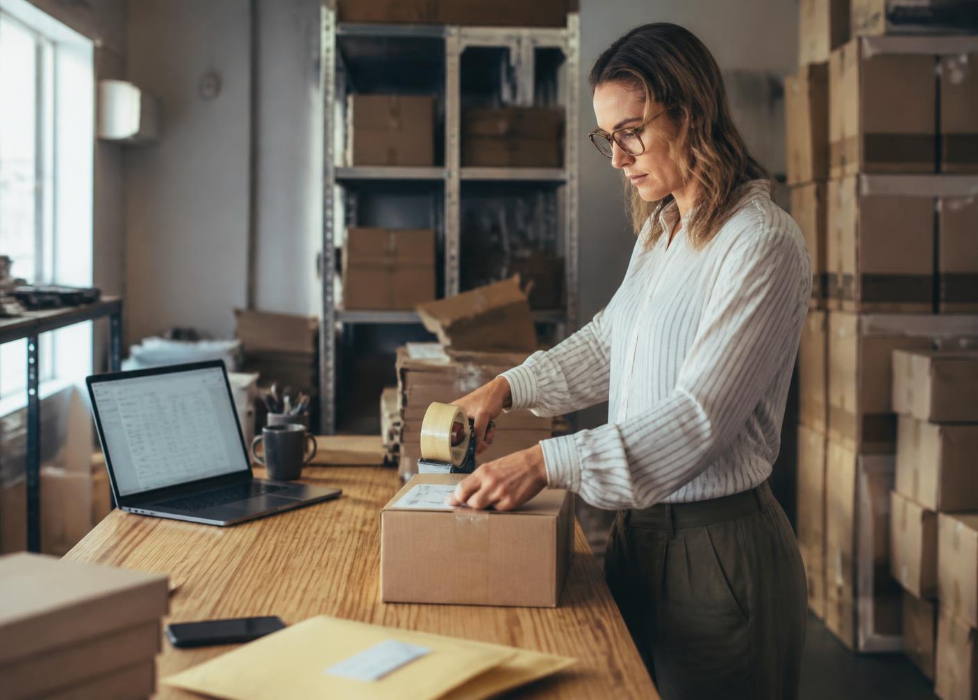 Online store owner packing customer orders beside a laptop showing store performance